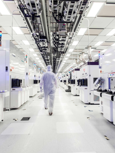 A technician in a white cleanroom suit walks through a semiconductor fabrication facility lined with manufacturing equipment.